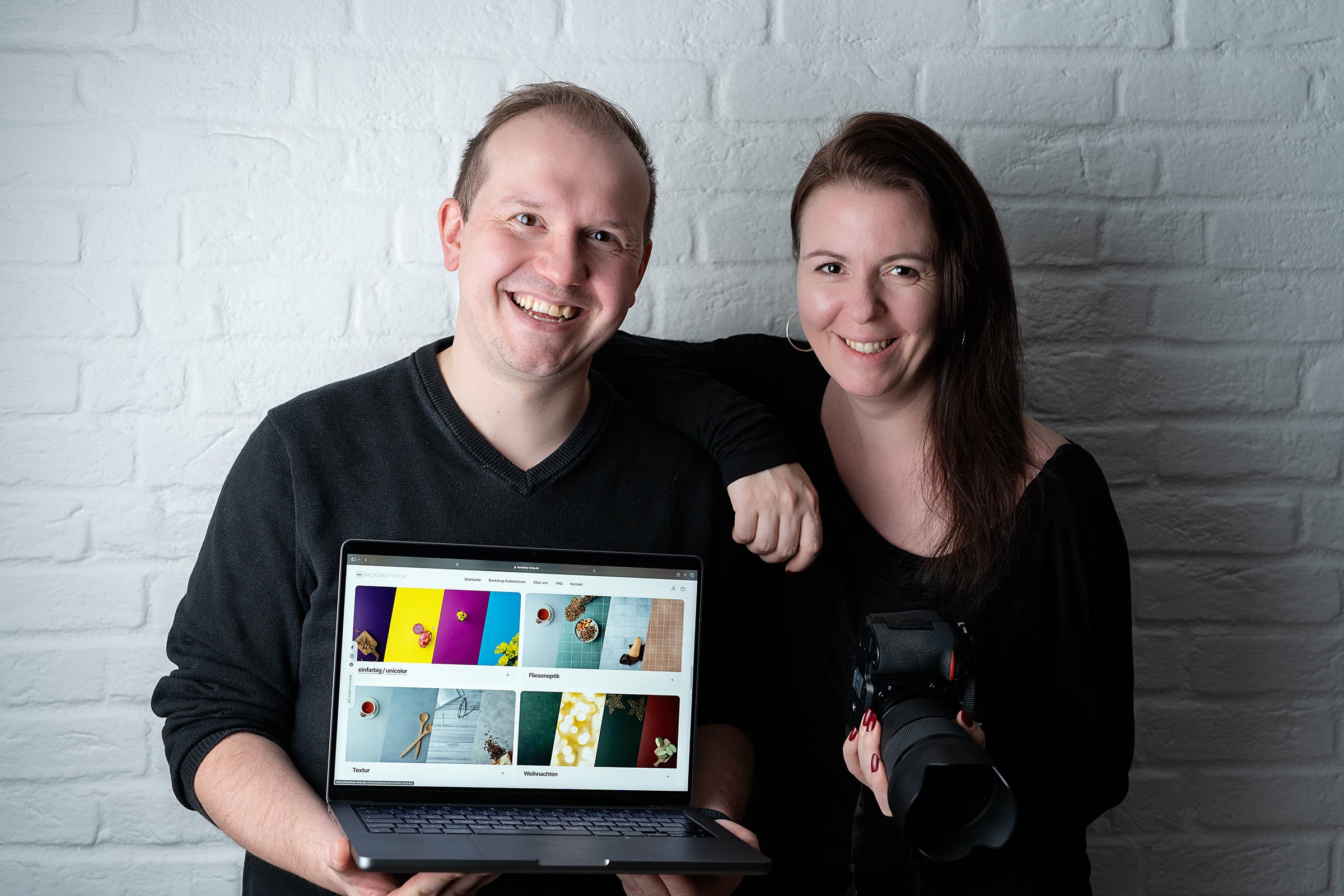 Katja und David, die Gründer des Backdrop Shops, vor einer weißen Ziegelwand. David präsentiert einen Laptop mit der Website des Shops, während Katja eine Kamera hält – das Team hinter kreativen Fotohintergründen.