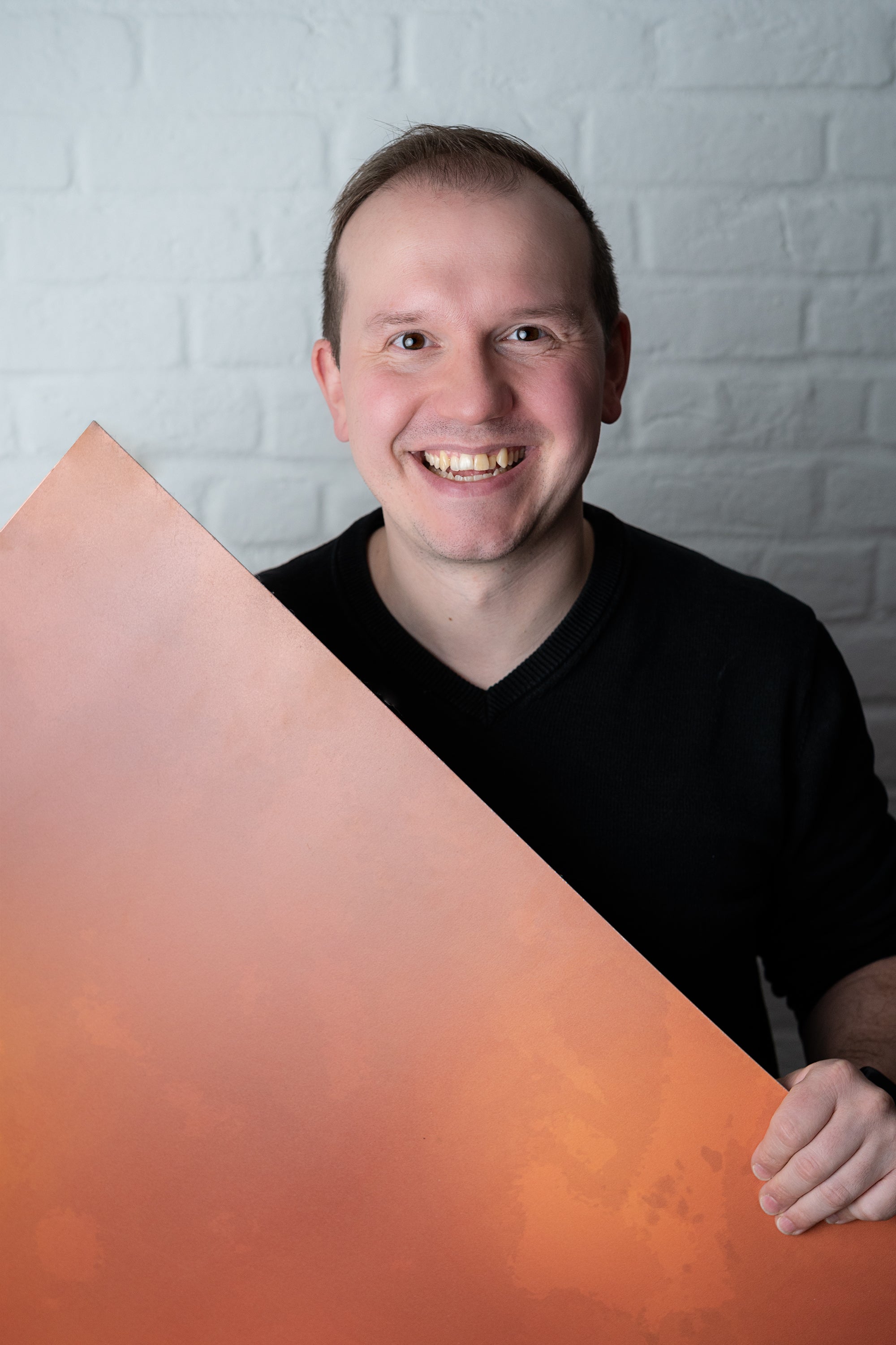 David, Mitgründer des Backdrop Shops, vor einer weißen Ziegelwand. Er hält einen orangefarbenen Fotohintergrund in den Händen und lächelt – der Experte für Technik und Logistik hinter den Backdrops.
