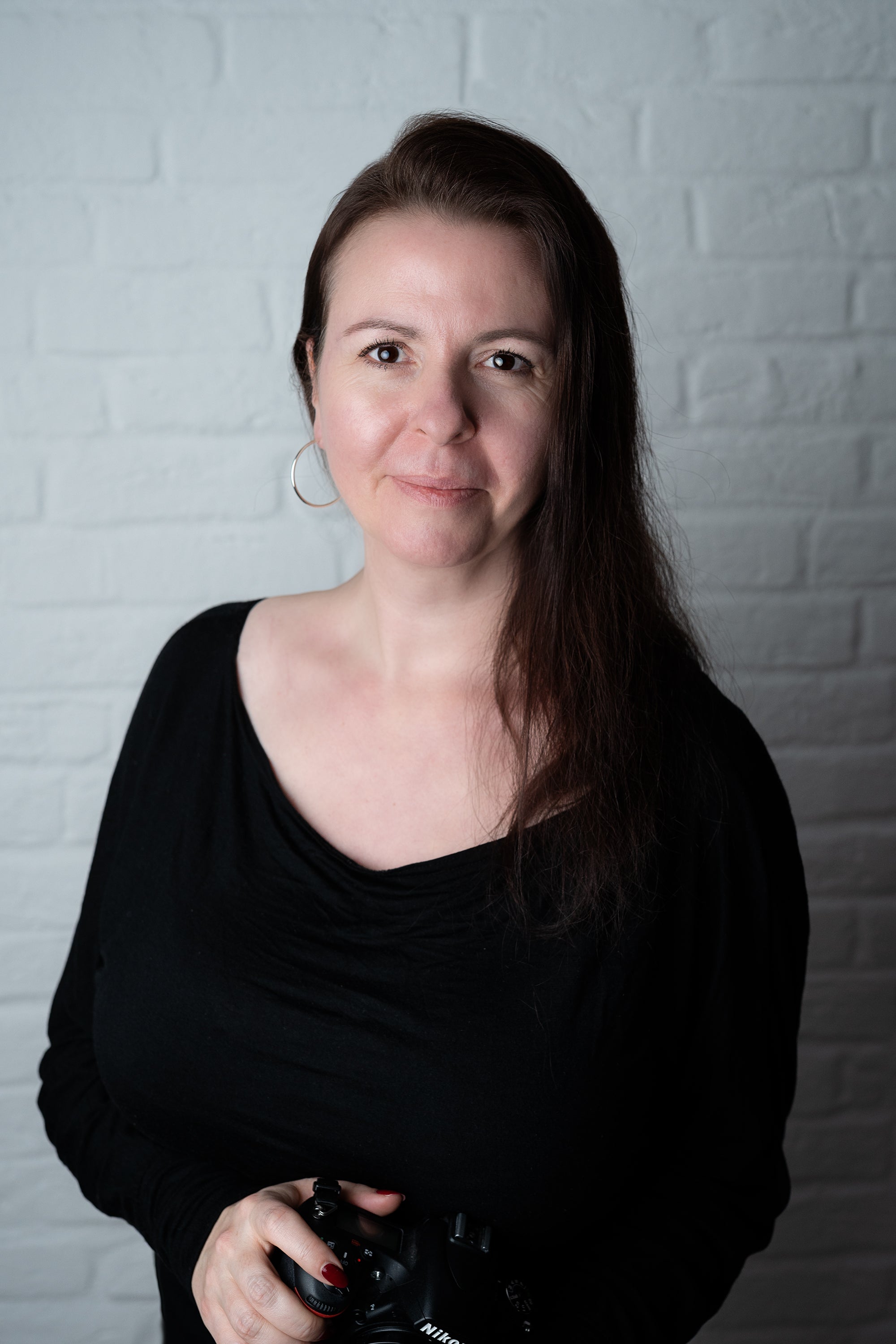 Katja, Gründerin des Backdrop Shops, vor einer weißen Ziegelwand. Sie trägt schwarze Kleidung und hält eine Kamera in den Händen – die kreative Persönlichkeit hinter den Fotohintergründen.