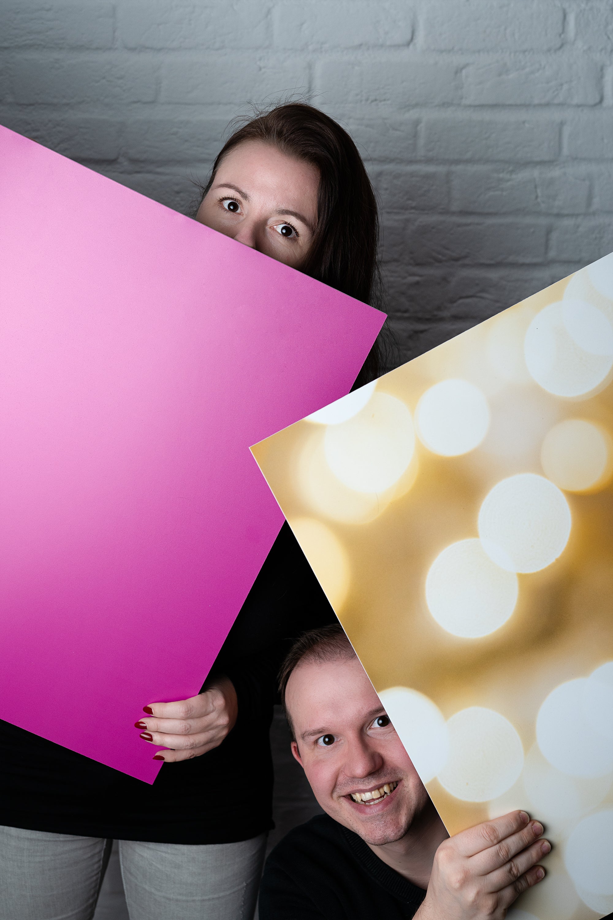 Katja und David, die Gründer des Backdrop Shops, posieren spielerisch hinter zwei Fotohintergründen. Katja hält einen pinken Backdrop, während David hinter einem goldenen Backdrop mit Bokeh-Effekt hervorblickt. Perfekt für kreative Fotografie.