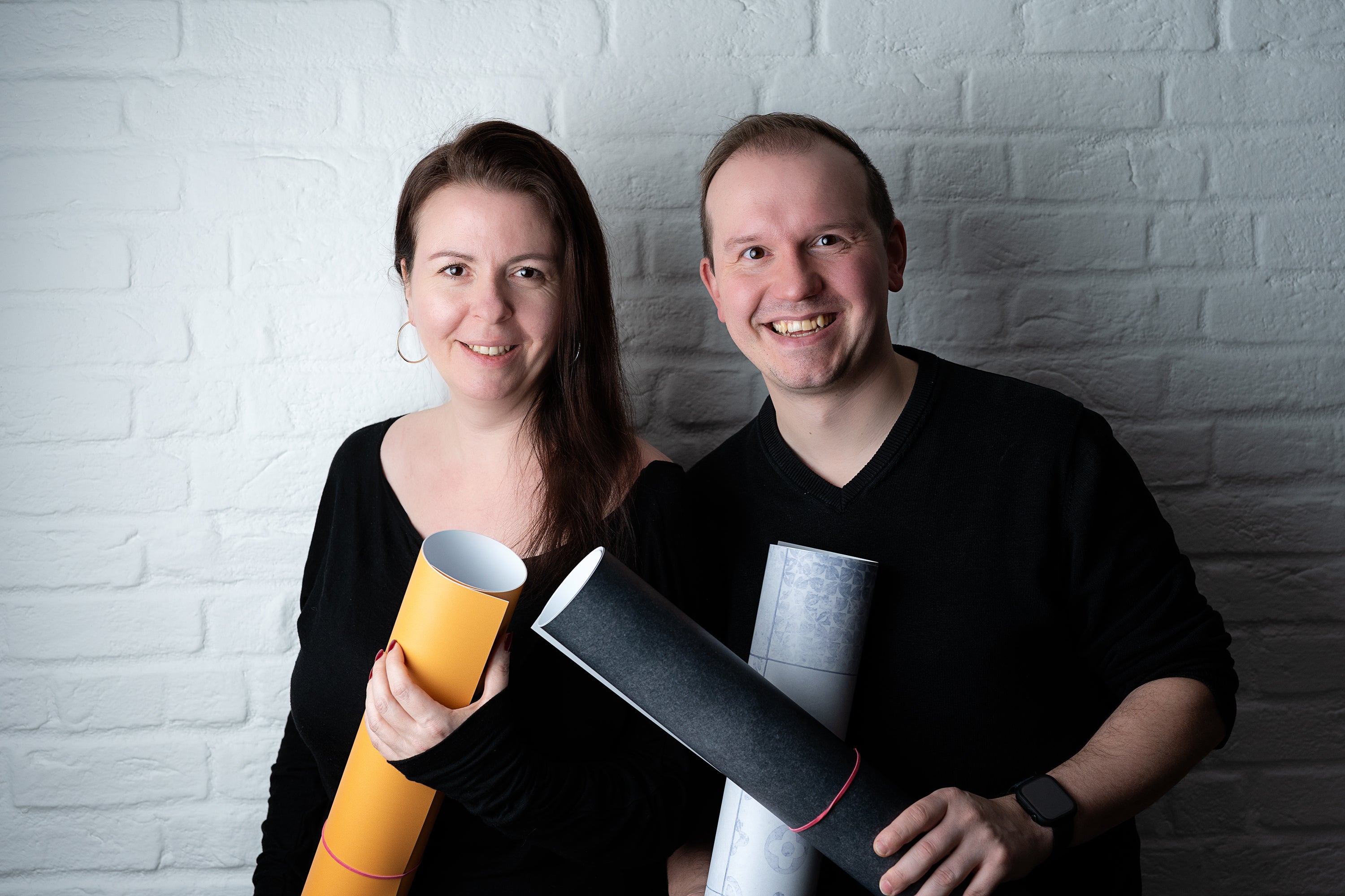 Katja und David, die Gründer des Backdrop Shops, vor einer weißen Ziegelwand. Beide lächeln und halten zusammengerollte Fotohintergründe in den Händen – das kreative Team hinter den vielseitigen Backdrops.