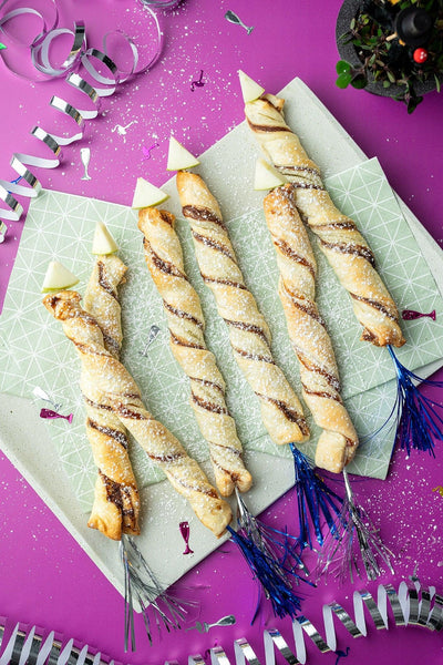 Silvester-Flatlay mit gedrehten Blätterteig-Raketen, Konfetti und Luftschlangen auf Backdrop „Granatapfel" – pinker Fotohintergrund