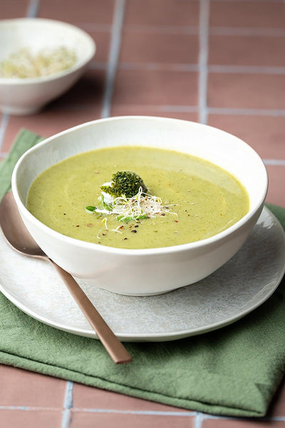 Brokkoli-Cremesuppe in weißer Schale mit Kupferlöffel auf grünem Leinentuch vor Backdrop „Hokkaido" – Terracotta Fliesenoptik-Fotohintergrund