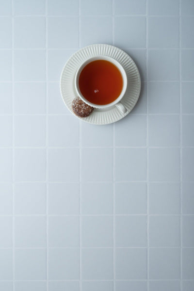 Top-down Teetasse mit Untertasse und Gebäck auf Backdrop „Kokosjoghurt" – cleaner weißer Fliesenoptik-Fotohintergrund