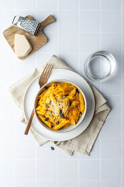 Top-down Kürbis-Penne mit Parmesan und Kürbiskernen auf Backdrop „Kokosjoghurt" – weißer Fliesenhintergrund mit Tischszene