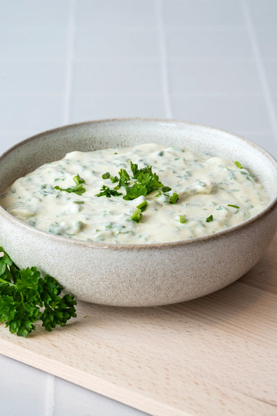 Kräuterquark in Keramikschüssel mit Petersilie auf Holzbrett vor Backdrop „Kokosjoghurt" – heller weißer Fliesenoptik-Fotohintergrund